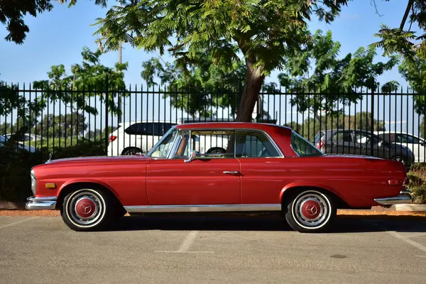 Caddeye güneş ışığı altında park edilmiş klasik kırmızı bir Mercedes Benz 280 SE Coupe. İkonik Alman lüks aracında krom bir ızgara, belirgin yuvarlak farlar ve 1960 'ların klasik tasarım detayları yer almaktadır..