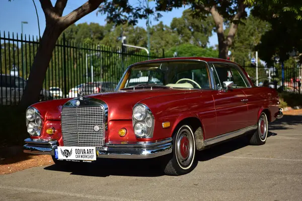 Caddeye güneş ışığı altında park edilmiş klasik kırmızı bir Mercedes Benz 280 SE Coupe. İkonik Alman lüks aracında krom bir ızgara, belirgin yuvarlak farlar ve 1960 'ların klasik tasarım detayları yer almaktadır..