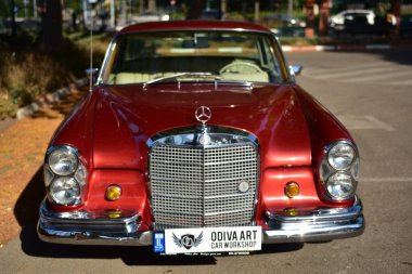 Caddeye güneş ışığı altında park edilmiş klasik kırmızı bir Mercedes Benz 280 SE Coupe. İkonik Alman lüks aracında krom bir ızgara, belirgin yuvarlak farlar ve 1960 'ların klasik tasarım detayları yer almaktadır..