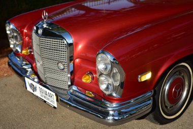 Caddeye güneş ışığı altında park edilmiş klasik kırmızı bir Mercedes Benz 280 SE Coupe. İkonik Alman lüks aracında krom bir ızgara, belirgin yuvarlak farlar ve 1960 'ların klasik tasarım detayları yer almaktadır..