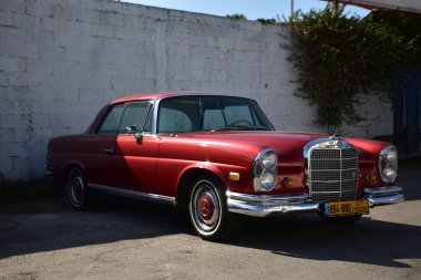 Caddeye güneş ışığı altında park edilmiş klasik kırmızı bir Mercedes Benz 280 SE Coupe. İkonik Alman lüks aracında krom bir ızgara, belirgin yuvarlak farlar ve 1960 'ların klasik tasarım detayları yer almaktadır..
