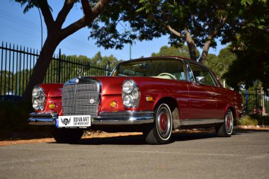 Caddeye güneş ışığı altında park edilmiş klasik kırmızı bir Mercedes Benz 280 SE Coupe. İkonik Alman lüks aracında krom bir ızgara, belirgin yuvarlak farlar ve 1960 'ların klasik tasarım detayları yer almaktadır..