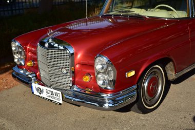 Caddeye güneş ışığı altında park edilmiş klasik kırmızı bir Mercedes Benz 280 SE Coupe. İkonik Alman lüks aracında krom bir ızgara, belirgin yuvarlak farlar ve 1960 'ların klasik tasarım detayları yer almaktadır..