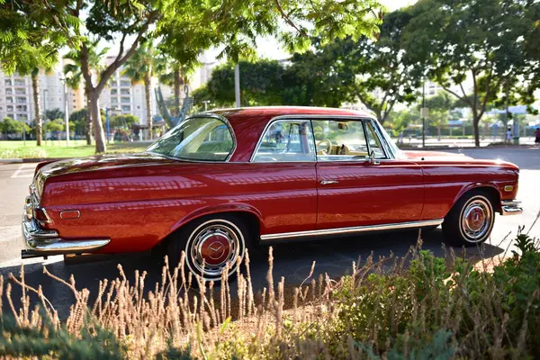 Caddeye güneş ışığı altında park edilmiş klasik kırmızı bir Mercedes Benz 280 SE Coupe. İkonik Alman lüks aracında krom bir ızgara, belirgin yuvarlak farlar ve 1960 'ların klasik tasarım detayları yer almaktadır..