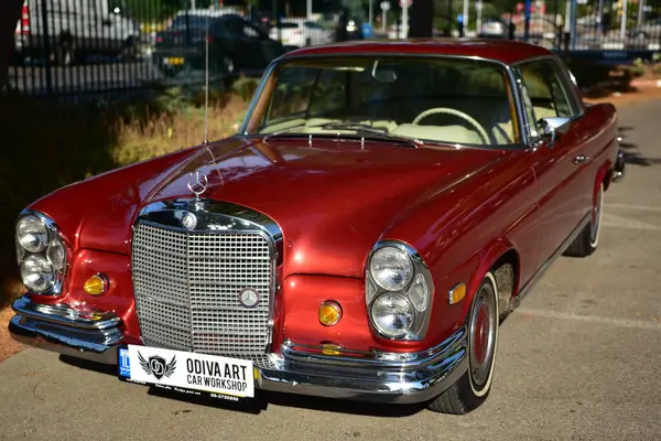Caddeye güneş ışığı altında park edilmiş klasik kırmızı bir Mercedes Benz 280 SE Coupe. İkonik Alman lüks aracında krom bir ızgara, belirgin yuvarlak farlar ve 1960 'ların klasik tasarım detayları yer almaktadır..