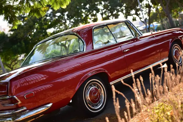 Caddeye güneş ışığı altında park edilmiş klasik kırmızı bir Mercedes Benz 280 SE Coupe. İkonik Alman lüks aracında krom bir ızgara, belirgin yuvarlak farlar ve 1960 'ların klasik tasarım detayları yer almaktadır..