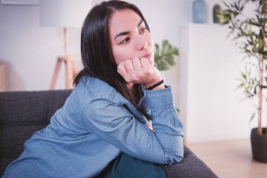 Evindeki kanepede oturan genç bir kadın yüzünde hüzünlü ve düşünceli bir ifadeyle.