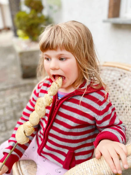 小さな幼児の女の子を保持串や棒にねじれスティックパン 火の炎の上にロースト ドイツで人気のパーティーやキャンプバーベキューの食べ物を噛んだり食べたりする面白い幸せな子供はStockbrotと呼ばれます — ストック写真