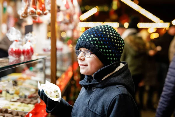 男の子子供かわいい子がバナナを食べるチョコレート Marshmellows ジンジャーブレッド ナッツと甘いスタンド近くのカラフルな振りかけるだらけ ドイツのクリスマス マーケットに幸せな少年 — ストック写真