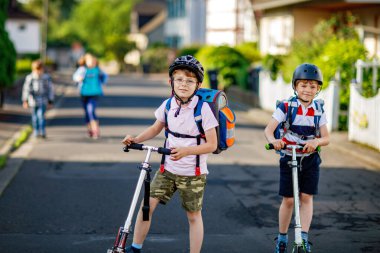 Güneşli bir günde, ellerinde scooterla şehirde gezen kasklı iki okul çocuğu. Renkli giysiler içinde okula giderken bisiklet süren mutlu çocuklar. Çocuklar için güvenli okul yolu..