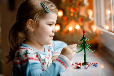 Küçük kız pencerenin kenarında oturuyor ve küçük cam Noel ağacını küçük Noel oyuncaklarıyla süslüyor. Mutlu sağlıklı çocuk geleneksel aile bayramını kutluyor. Sevimli bebek