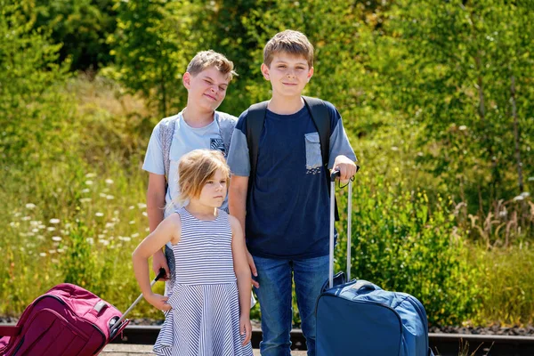 Three children, school boys and preschool girl with suitcases before leaving for summer vacation camp. Happy kids, siblings, twins brothers going on journey, family road trip waiting for train