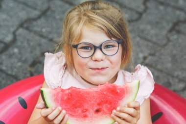 Cute little girl with glasses eating watermelon on inflatable ring in summertime. Happy smiling preschool child having fun. Healthy summer food and snacks for kids