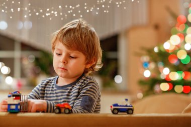 Evde arabalarla ve oyuncaklarla oynayan küçük sarışın çocuk. Anaokulunun çocuğu eğleniyor. Arka planda Noel ağacı