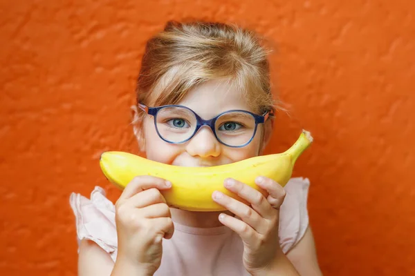 stock image Happy little child girl with yellow banana like smile on orange background. Preschool girl with glasses smiling. Healthy fruits for children.