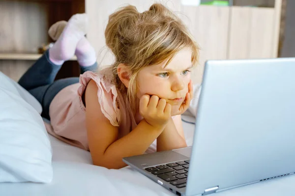 Preschool girl watching videos on laptop, notebook,in bed on clean white linens. Indoors activity with children. Freelance, distance learning or work from home with kids concept. Happy child.