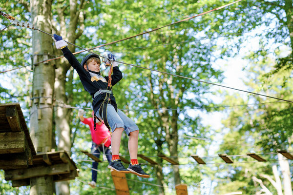 Two children in forest adventure park. Kids boys in helmet climbs on high rope trail. Agility skills and climbing outdoor amusement center for children. Outdoors activity for kid and families