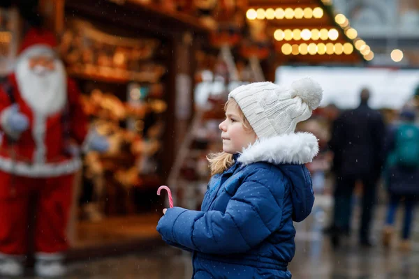 クリスマスマーケットのお菓子スタンドからのお菓子杖を持つ小さなかわいい就学前の女の子 ドイツの伝統的な家庭市場で幸せな子供 雪の中でカラフルな冬服の就学前 — ストック写真