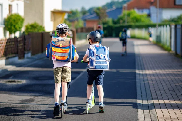 在阳光明媚的城市 两个戴着安全帽 背着背包骑摩托车的小学生 穿着五颜六色衣服的快乐孩子们骑自行车去上学 儿童在户外上学的安全方法 — 图库照片