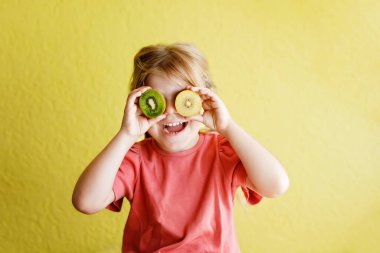 Happy little child girl with yellow and green kiwi fruits on yellow background. Preschool girl smiling. Healthy fruits for children.