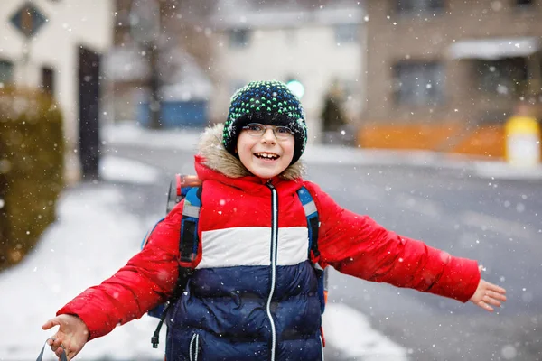 小学生の少年は雪の中学校に歩いている 楽しさと最初の雪で遊んで幸せな子供 学生でアイグラスバックパックでカラフルな冬服 — ストック写真