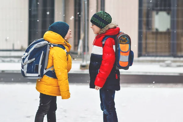 雪の中 小学生の男の子2人が学校に歩いています 楽しい時間を過ごし 初めての雪で遊ぶ幸せな子供たち 兄弟やカラフルな冬服のバックパックと親友 — ストック写真