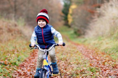 Sonbahar Ormanı Parkı 'nda renkli, sıcak giysiler içinde bisiklet süren küçük çocuk. Doğada güneşli bir sonbahar gününde bisiklet süren aktif bir çocuk. Güvenlik, spor, çocuk konseptiyle eğlence. Çocuklar için gündelik moda