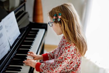 Little happy girl playing piano in living room. Cute preschool child with eye glasses having fun with learning to play music instrument