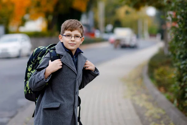 Happy kid boy with glasses and backpack or satchel. Schoolkid in stylish fashon coan on the way to middle or high school on cold autumn day. Healthy child outdoors on the street, on rainy day