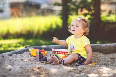 Happy toddler girl playing in sand on outdoor playground. Baby having fun on sunny warm summer sunny day. Active child with sand toys and in colorful fashion clothes