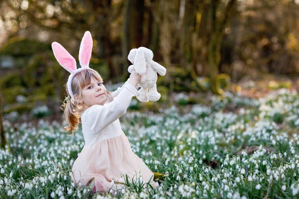 可爱的小女孩 带着复活节兔子的耳朵 在阳光灿烂的春天的森林里抱着柔软的毛绒玩具 在户外 带着许多雪花的可爱的快乐的孩子 基督教节日的概念 — 图库照片
