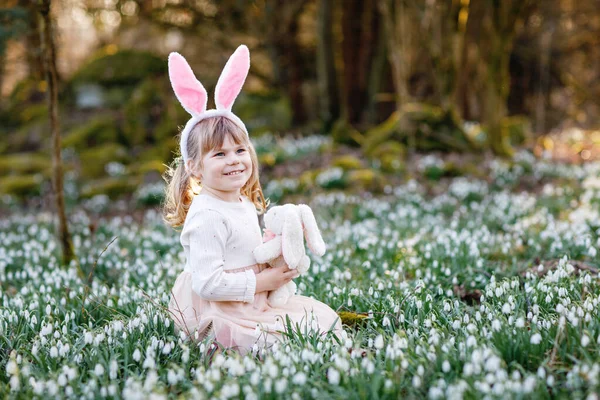 可爱的小女孩 带着复活节兔子的耳朵 在阳光灿烂的春天的森林里抱着柔软的毛绒玩具 在户外 带着许多雪花的可爱的快乐的孩子 基督教节日的概念 — 图库照片