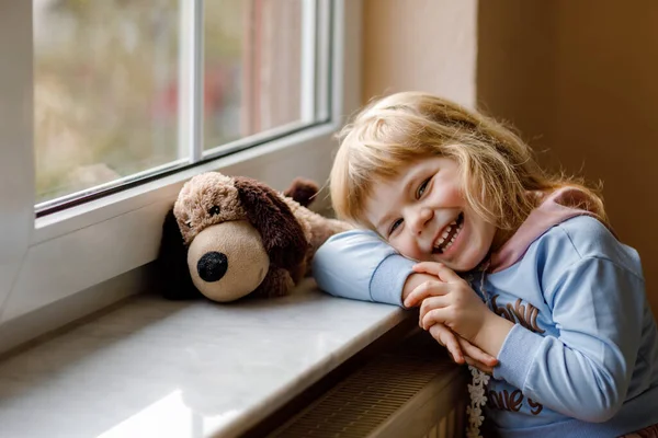かわいい幼児の女の子が窓のそばに座って外を見ています 柔らかい犬のおもちゃで子供の夢を見て幸せな気分 孤独な子 — ストック写真
