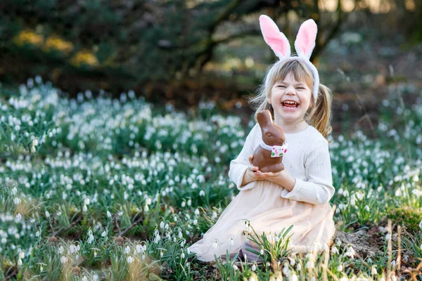 快乐的小女孩 带着复活节兔子的耳朵 在阳光明媚的春天的森林里吃着巧克力 可爱的孩子 有很多雪花 基督教节日的概念 — 图库照片