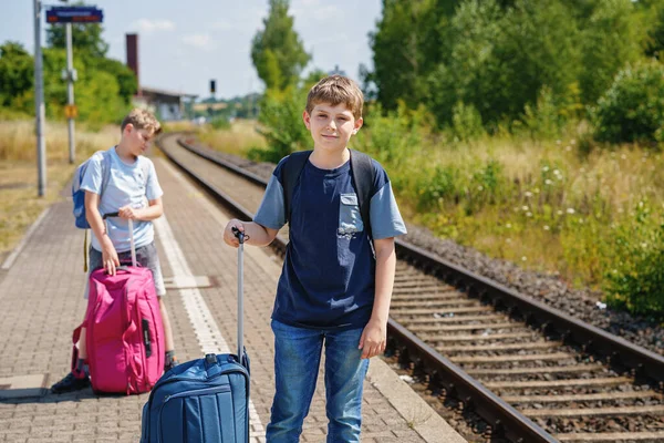 Two children, school boys with suitcases before leaving for summer vacation camp. Happy kids, siblings, twins brothers going on journey, family road trip waiting for train