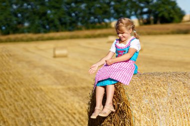 Cute little kid girl in traditional Bavarian costume in wheat field. Happy child with hay bale during Oktoberfest in Munich. Preschool girl play at hay bales during summer harvest time in Germany