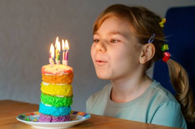 Happy little preschool girl celebrating birthday. Closeup of child with homemade rainbow cake, indoor. Happy healthy toddler blowing six candles on cake. Selective focus on cake.