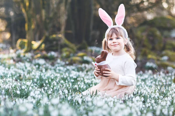 快乐的小女孩 带着复活节兔子的耳朵 在阳光明媚的春天的森林里吃着巧克力 可爱的孩子 有很多雪花 基督教节日的概念 — 图库照片