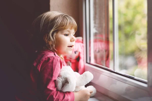 かわいい幼児の女の子は窓のそばに座って雨の日を見ています 人形と柔らかいおもちゃの幸せな気分で子供を夢見ています コロナウイルスパンデミック時の自己分離概念 孤独な子 — ストック写真