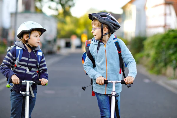 晴れた日にバックパックで街のスクーターに乗って安全ヘルメットに乗っている2人の子供の男の子 学校に行く途中でカラフルな服を自転車で幸せな子供たち 学校への屋外の子供のための安全な方法 — ストック写真