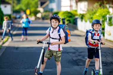 Güneşli bir günde, ellerinde scooterla şehirde gezen kasklı iki okul çocuğu. Renkli giysiler içinde okula giderken bisiklet süren mutlu çocuklar. Çocuklar için güvenli okul yolu..
