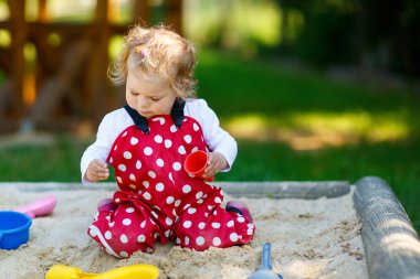 Dışarıda parkta kumlarda oynayan tatlı bir kız çocuğu. Kırmızı sakız pantolonlu güzel bebek güneşli yaz gününde eğleniyor. Renkli kum oyuncakları olan çocuk. Sağlıklı aktif bebek dışarıda oyunlar oynuyor.