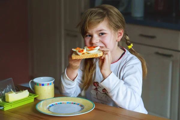 小さな笑顔の女の子は家で朝食を持っています 就学前の子供はゆで卵でサンドイッチを食べる 幸せな子供たち 健康的な食べ物と食事 — ストック写真
