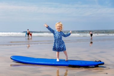 Little cute toddler girl at the Ballybunion surfer beach, having fun on surfboard for the first time, west coast of Ireland. Happy child enjoying Irish summer with family clipart