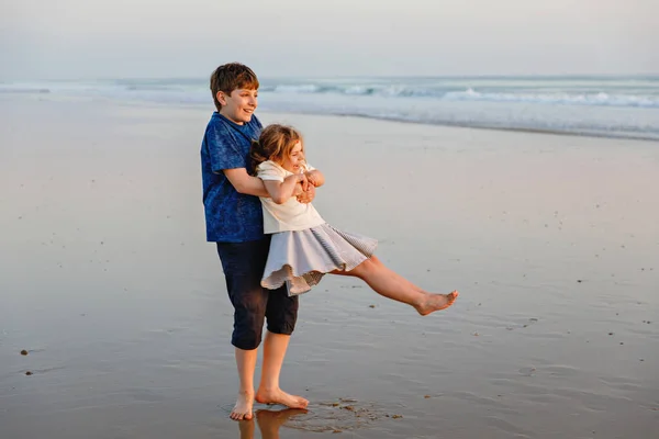 海のビーチで2人の子供 10代の男の子と小さな就学前の妹は日没までに楽しんでいます 家族のための海の休暇 兄弟は笑って笑っています 幸せと愛 — ストック写真