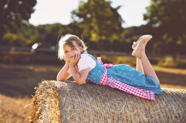 Cute little kid girl in traditional Bavarian costume in wheat field. Happy child with hay bale during Oktoberfest in Munich. Preschool girl play at hay bales during summer harvest time in Germany