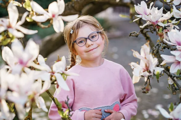 Cute spring fashion preschool girl with glasses under blossom magnolia tree. Little happy child and spring