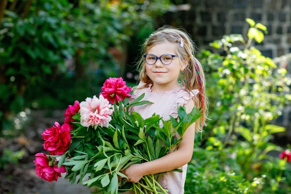 赤とピンクの牡丹の花の巨大な花束を持つかわいい愛らしい小さな就学前の女の子 暖かい春や夏の日に家庭菜園で笑顔の就学前の子供の肖像画 — ストック写真