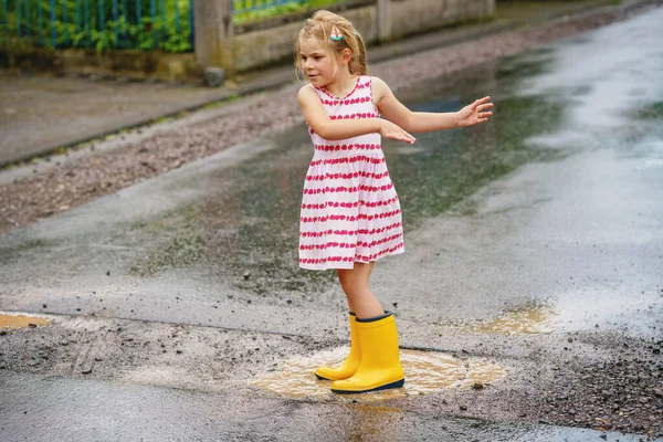 黄色のレインブーツを着用し 水たまりの間に歩いて幸せな小さな就学前の女の子 カラフルな服のかわいい子供が水たまりに飛び込み 水で飛び散ったり 屋外活動 — ストック写真