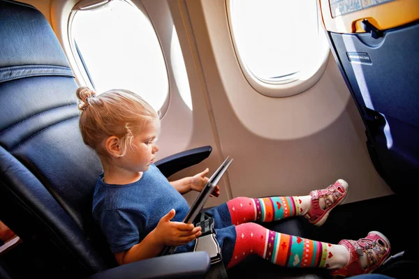 Little toddler girl traveling by plane. Small happy child sitting by aircraft window and using a digital tablet during the flight. Traveling abroad with kids
